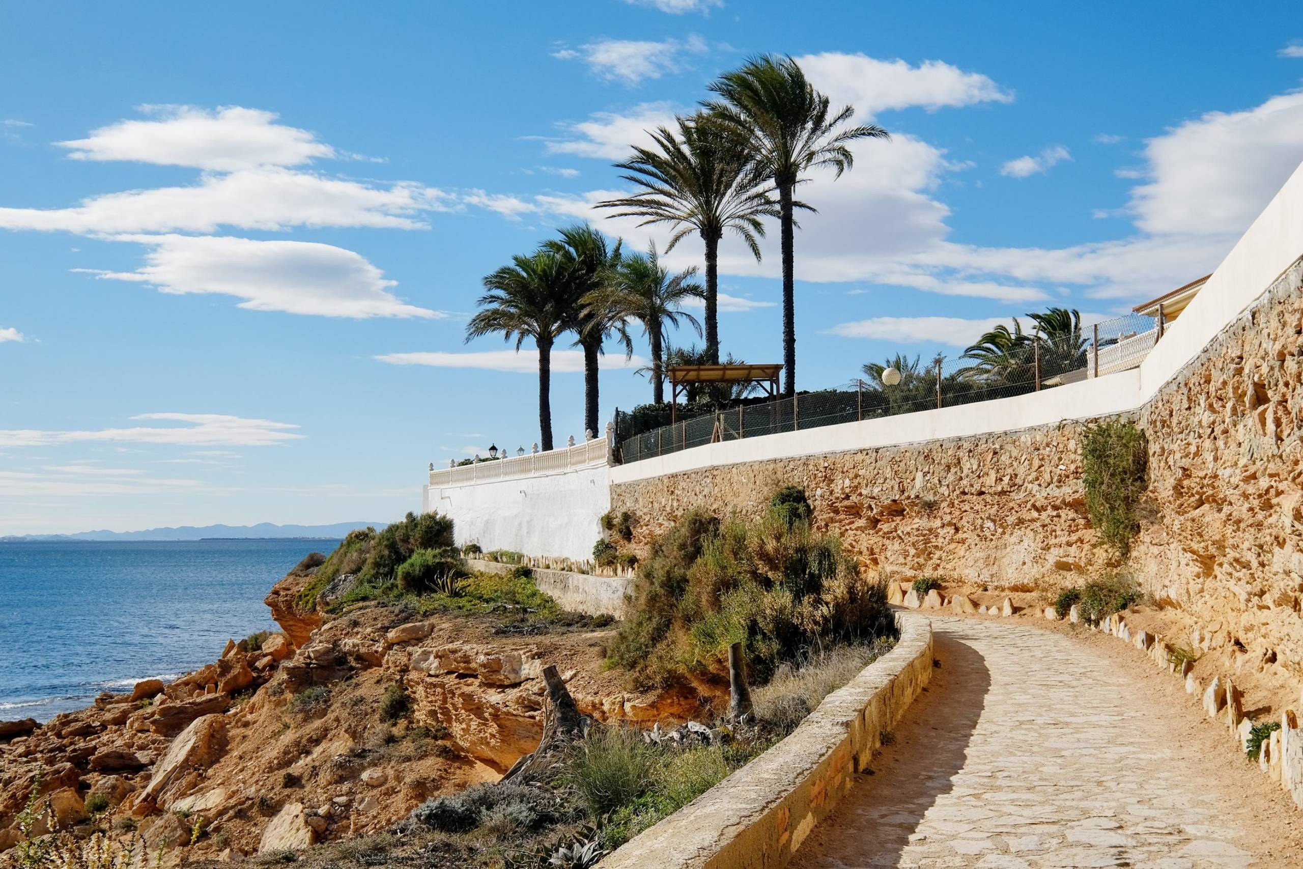 A stone pathway winds along a rocky cliff overlooking the blue sea, lined with palm trees under a cloudy sky.