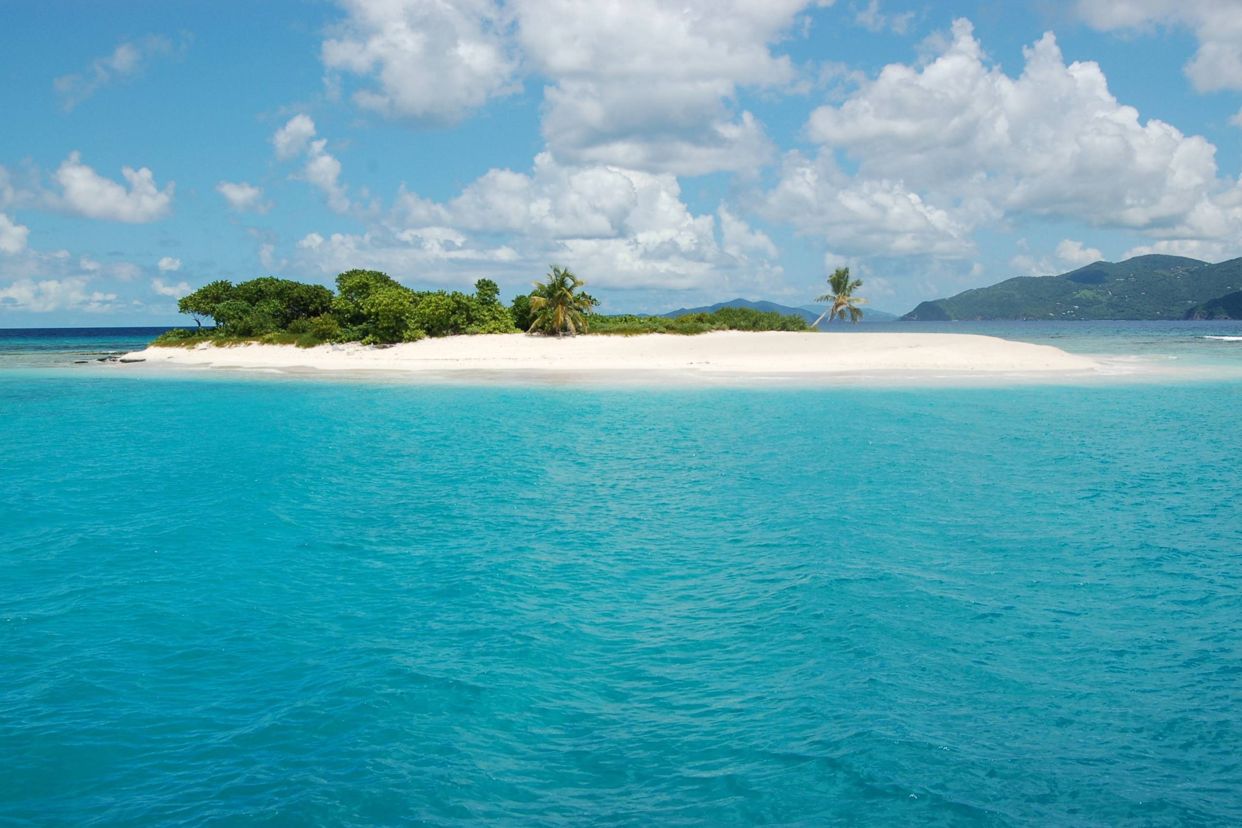 A pristine white sandbar dotted with palm trees emerges from the vibrant turquoise waters under a cloudy blue sky.