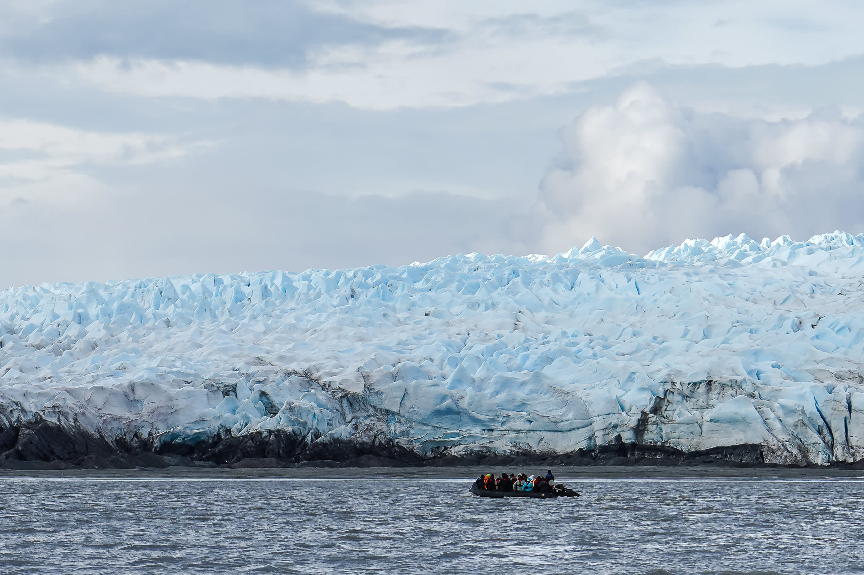 A small boat with people navigates choppy water in front of a massive, icy blue glacier under a cloudy sky.