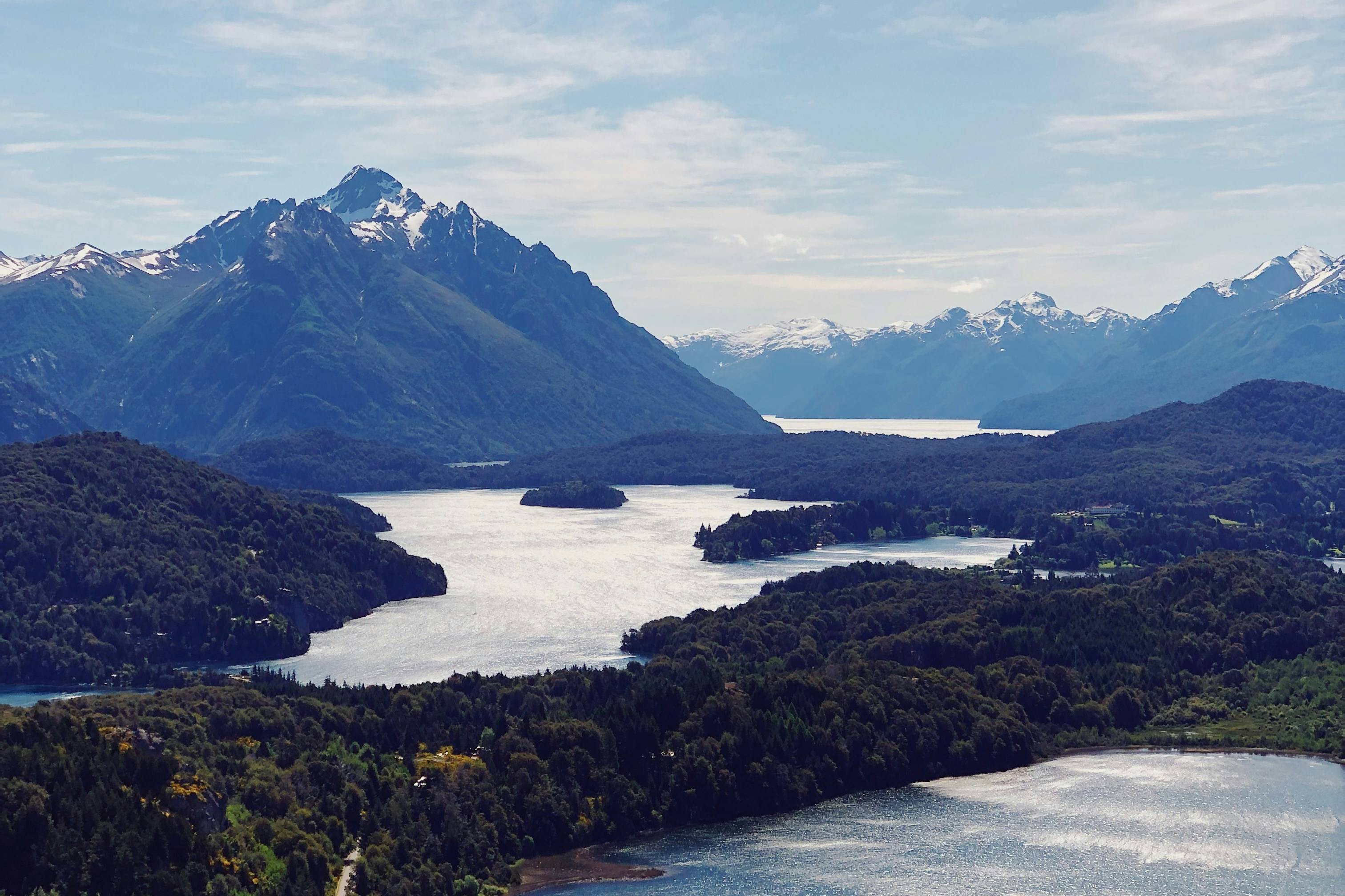 Lush green trees in the foreground lead to a serene lake and majestic snow-capped mountains under a cloudy blue sky in the background.