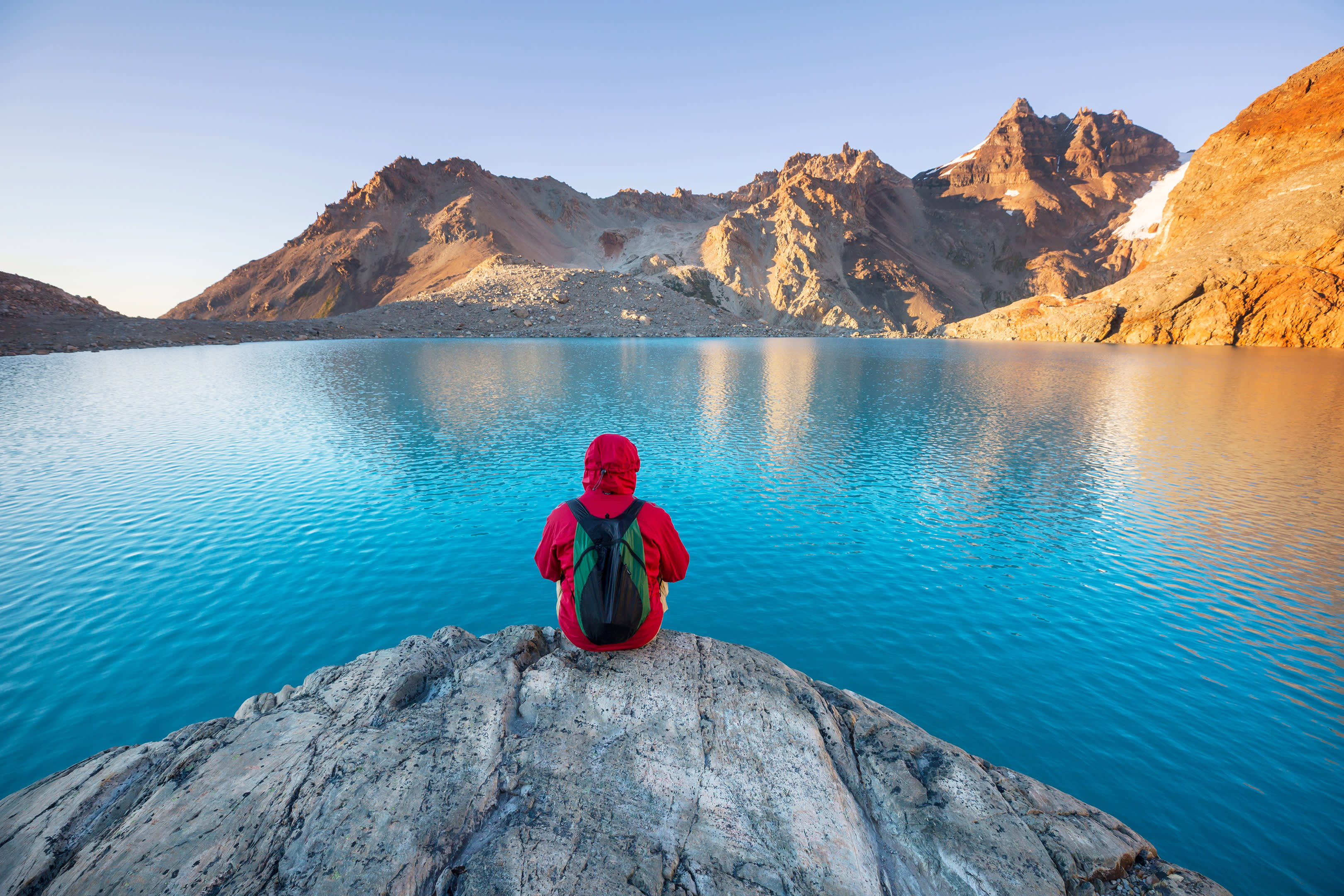 A lone hiker in a red jacket contemplates the serene turquoise waters of a mountain lake at sunrise.