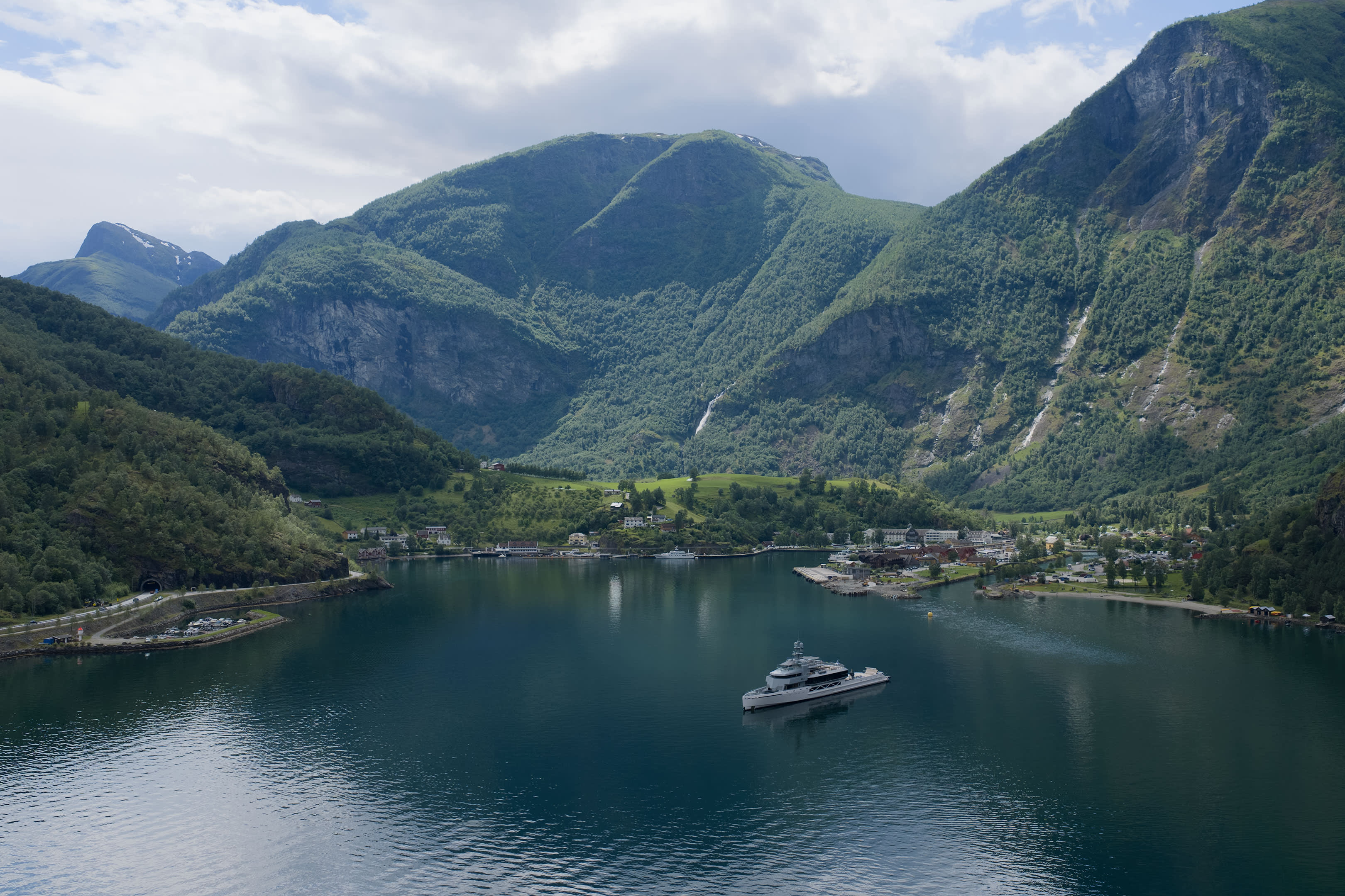 A sleek yacht cruises on a calm, dark blue fjord, with a small village nestled at the base of lush, green mountains in the background.