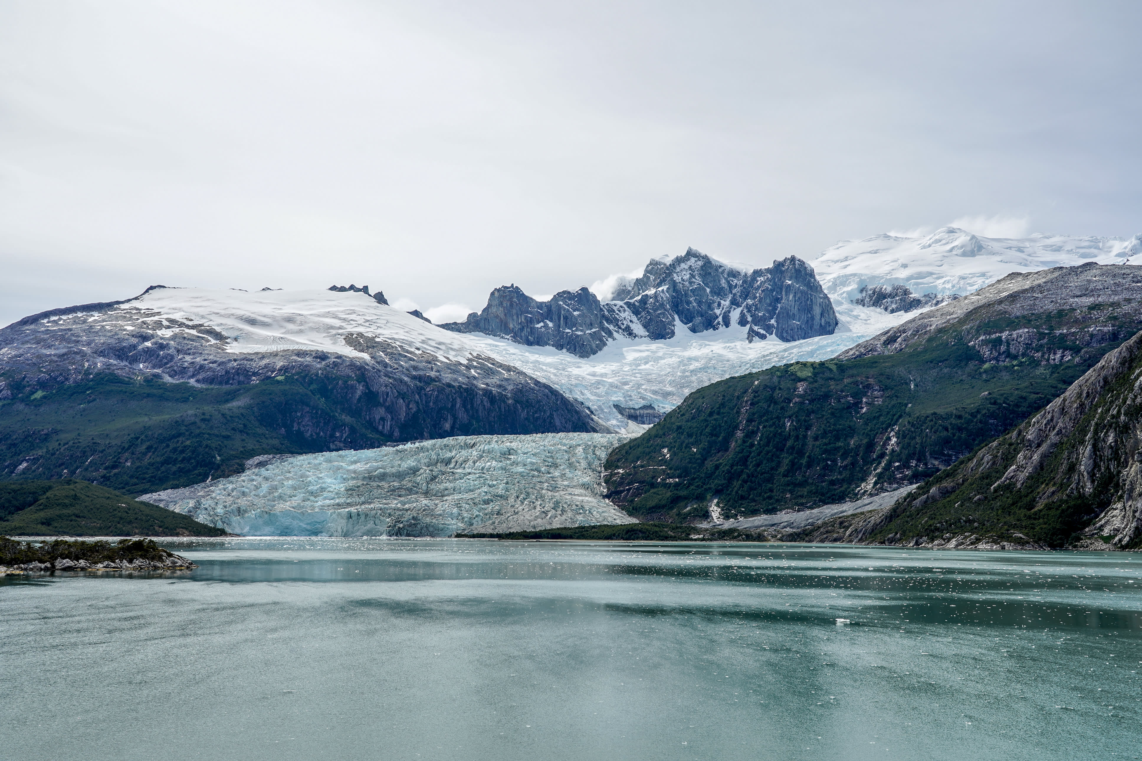 A vast, icy glacier flows down a mountain range into a calm, turquoise body of water.