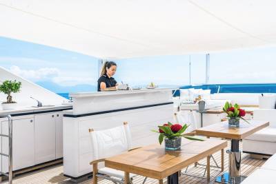 A woman in uniform prepares drinks and snacks at a sleek bar on a sun-drenched yacht deck.
