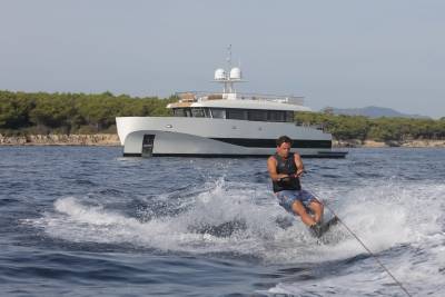 A man wakeboards through choppy blue water with a sleek white yacht anchored offshore.
