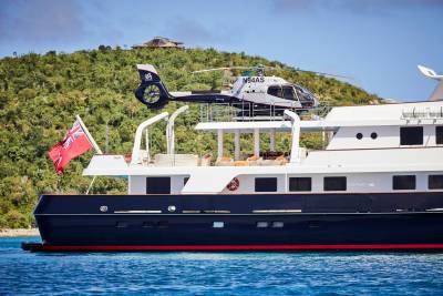 A luxurious yacht with a helicopter on deck cruises through clear blue waters near a lush green island.