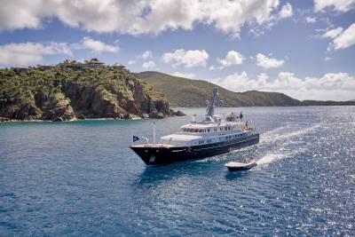 A luxurious yacht cruises through sparkling blue waters near a verdant, cliffside estate under a bright, cloudy sky.