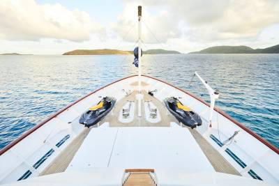 Two jet skis are secured on the bow of a luxurious yacht sailing through calm blue waters near a tropical island.