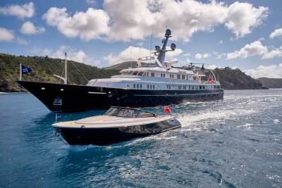 A sleek tender navigates the sparkling blue waters ahead of a majestic superyacht anchored near a lush, green island.