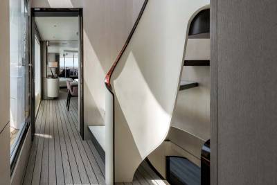 A modern staircase with a flowing white railing curves elegantly beside a sun-drenched hallway with wooden floors.