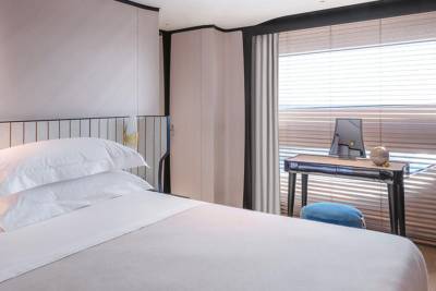 A modern bedroom features a plush bed, a sleek desk with a globe and monitor, and large windows with blinds.