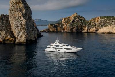 A luxurious white yacht cruises through calm blue waters near dramatic rocky cliffs under a partly cloudy sky.
