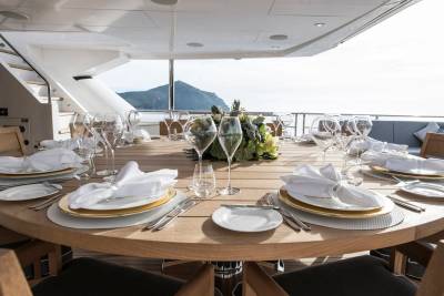 A beautifully set outdoor dining table on a yacht awaits guests with a stunning ocean and mountain backdrop.