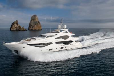A sleek white yacht cuts through the deep blue sea, leaving a foamy wake near dramatic rock formations.