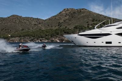 Two jet skiers race through the waves near a luxurious white yacht anchored off a rugged, tree-covered coastline.
