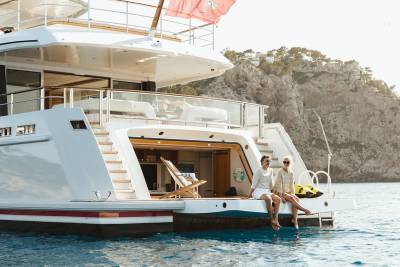 Two women relax on the stern of a luxurious yacht anchored near a rocky, tree-covered coastline.