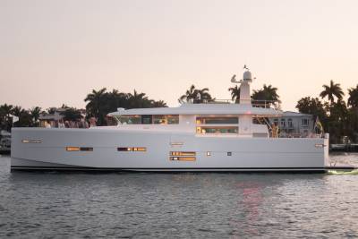 A sleek white yacht cruises through calm waters at dusk, its illuminated windows reflecting the warm glow of the setting sun.