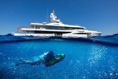 A person rides a sea scooter underwater near a luxurious yacht under a clear blue sky.