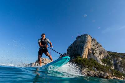 A man on a turquoise board glides through the water, creating a spray against a backdrop of rugged cliffs under a clear blue sky.