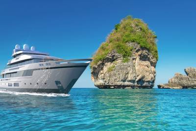 A sleek modern yacht glides through turquoise waters past a lush, rocky island under a clear blue sky.