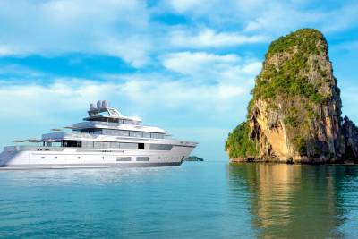 A sleek modern yacht cruises past a dramatic limestone karst island under a bright blue sky.