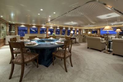 An elegant dining room aboard a yacht features a large, round table set for a formal meal, surrounded by plush seating and large windows overlooking the ocean.