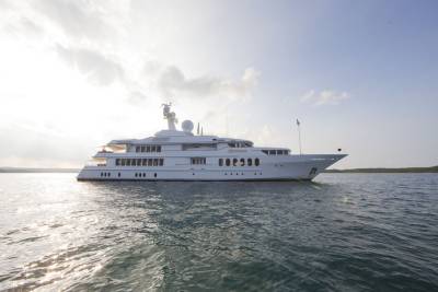 A gleaming white superyacht named "Huntress" glides serenely across calm waters under a bright, partly cloudy sky.