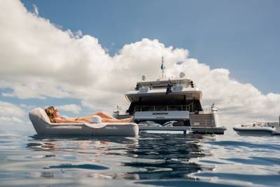 A woman relaxes on a floating lounger with a luxury yacht and a tender boat in the background under a cloudy sky.