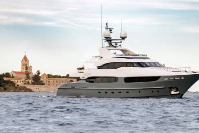 A sleek grey yacht cruises past a historic coastal monastery under a pale sky.