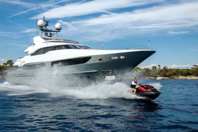 A man on a jet ski speeds through the water, kicking up a spray as a large yacht cruises by under a bright blue sky.