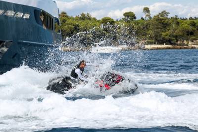 A man on a jet ski creates a dramatic splash as he speeds past a large yacht near a tree-lined coast.