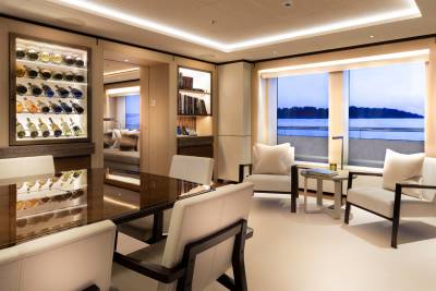 A well-lit interior features a wine cellar, dining table, and comfortable seating with a scenic water view.