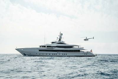 A sleek, modern yacht cruises through the choppy blue sea, accompanied by a small helicopter flying overhead.