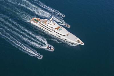 A luxurious superyacht cruises through deep blue waters, trailed by smaller speedboats and jet skis.