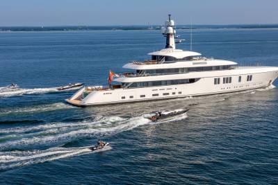 A massive white yacht cruises through the blue ocean, trailed by a jet ski and two speedboats.