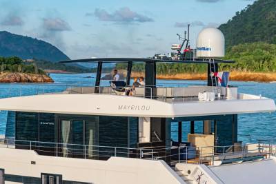 A sleek yacht named MAYRILOU cruises through calm blue waters near a lush, green coastline.