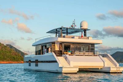 A luxurious catamaran glides through calm turquoise waters near a lush, green island under a soft, cloudy sky.