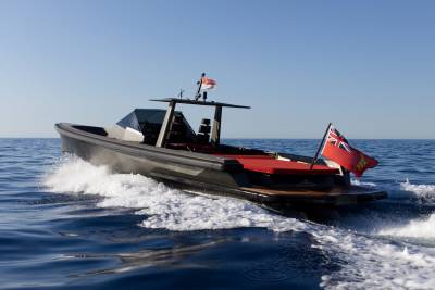 A sleek, dark boat cuts through the blue ocean waves under a clear sky, flying both the Indonesian and British flags.