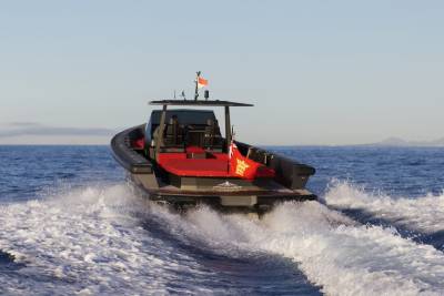 A sleek, dark boat with red accents cuts through the ocean, leaving a frothy white wake under a clear blue sky.