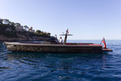 A man pilots a sleek, dark boat with red accents across the shimmering blue waters near a cliffside villa.