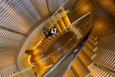 A woman carries a tray up a luminous, spiraling wooden staircase with a sleek glass railing.