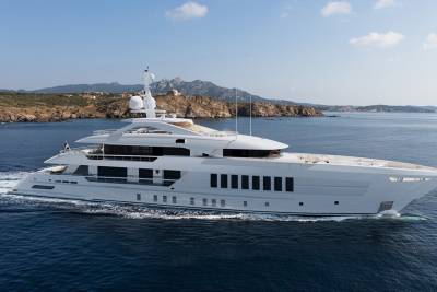 A luxurious white superyacht glides through the deep blue ocean near a rocky, verdant coastline under a clear sky.