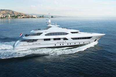A sleek white yacht glides through the azure waters, leaving a foamy wake against a backdrop of a coastal town.