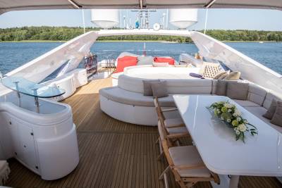 A luxurious yacht deck offers a serene setting with plush seating, a dining table, and a beautiful floral arrangement against a backdrop of blue water and lush green trees.