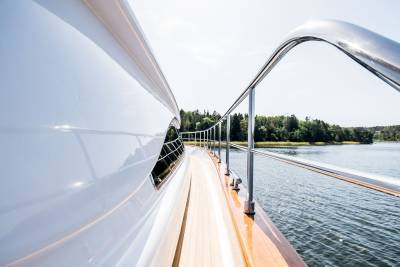 The gleaming white hull of a luxury yacht cuts through calm waters, bordered by a polished railing and lush green coastline.