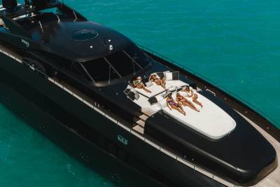 Five women in bikinis lounge on the sun deck of a sleek black yacht cruising through turquoise waters.