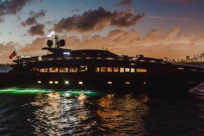 A luxurious yacht with green underwater lights glides through the water at dusk, with a city skyline in the background.