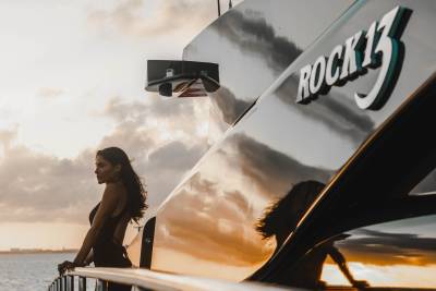 A woman in a black dress gazes out at the ocean from the deck of the yacht "ROCK 13" at sunset.