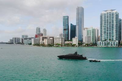 A sleek black yacht cruises through turquoise waters with a smaller boat in tow, against a backdrop of Miami's impressive skyline.