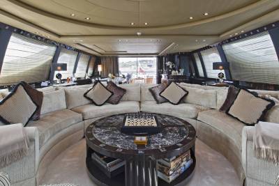 A luxurious yacht lounge features a circular sofa, marble-topped coffee table with a chessboard, and panoramic windows overlooking a scenic landscape.
